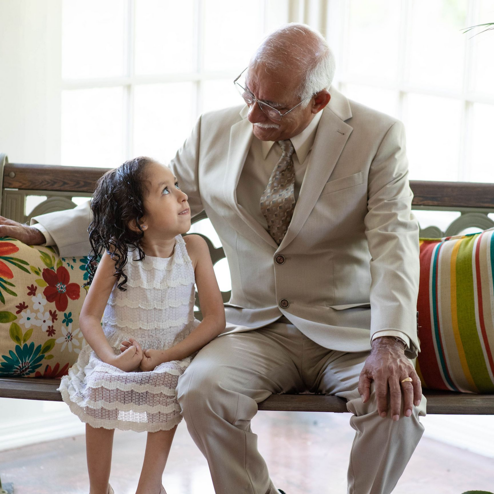 Elderly man in a beige suit smiling at a young girl on a couch in a bright living room.