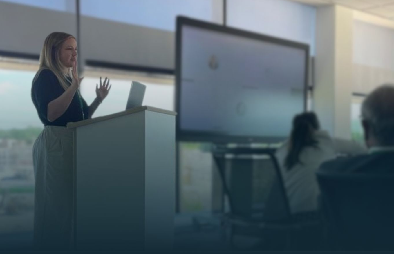 Woman giving a presentation at a podium, gesturing with hands. Large screen and audience visible in a bright office.