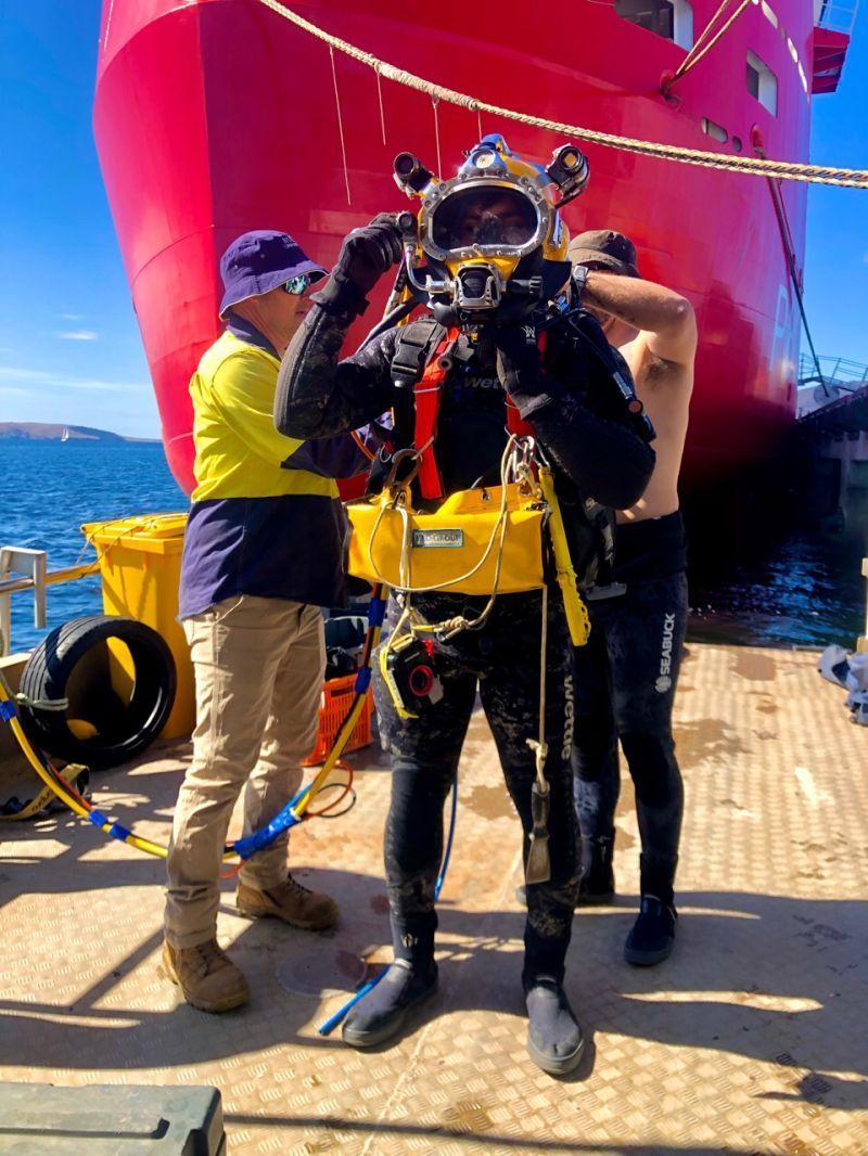 A diver in full gear prepares for an underwater mission, aided by two assistants on a dock near a red ship.