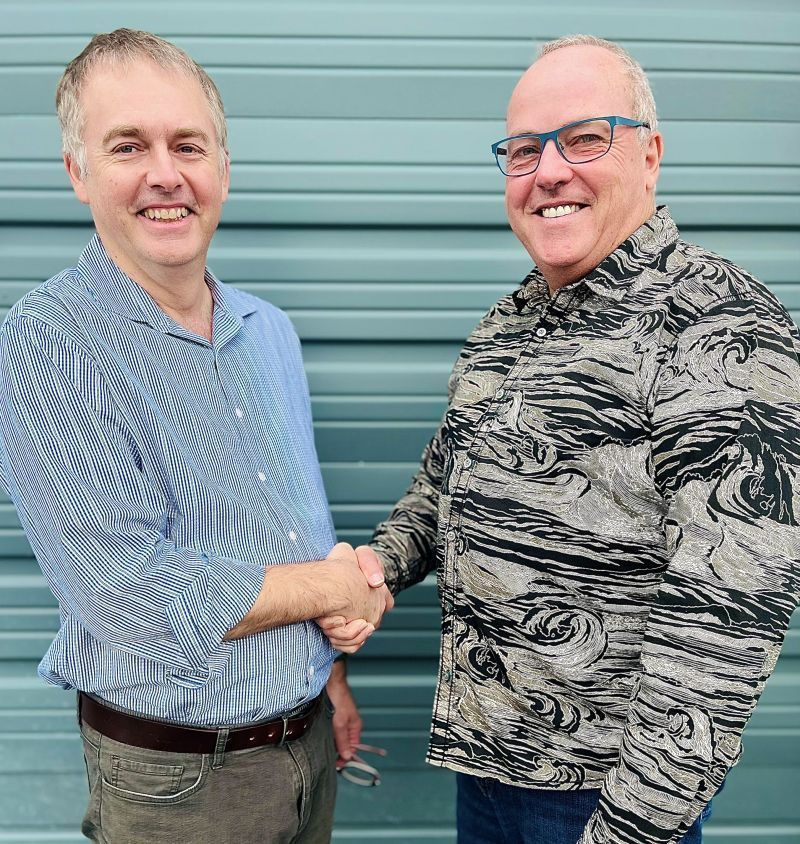 Two men shaking hands in front of a turquoise wall, smiling. One in a blue striped shirt, the other in a patterned shirt.