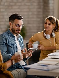 Three people at a table, two looking at a tablet, smiling, holding mugs. Office setting.