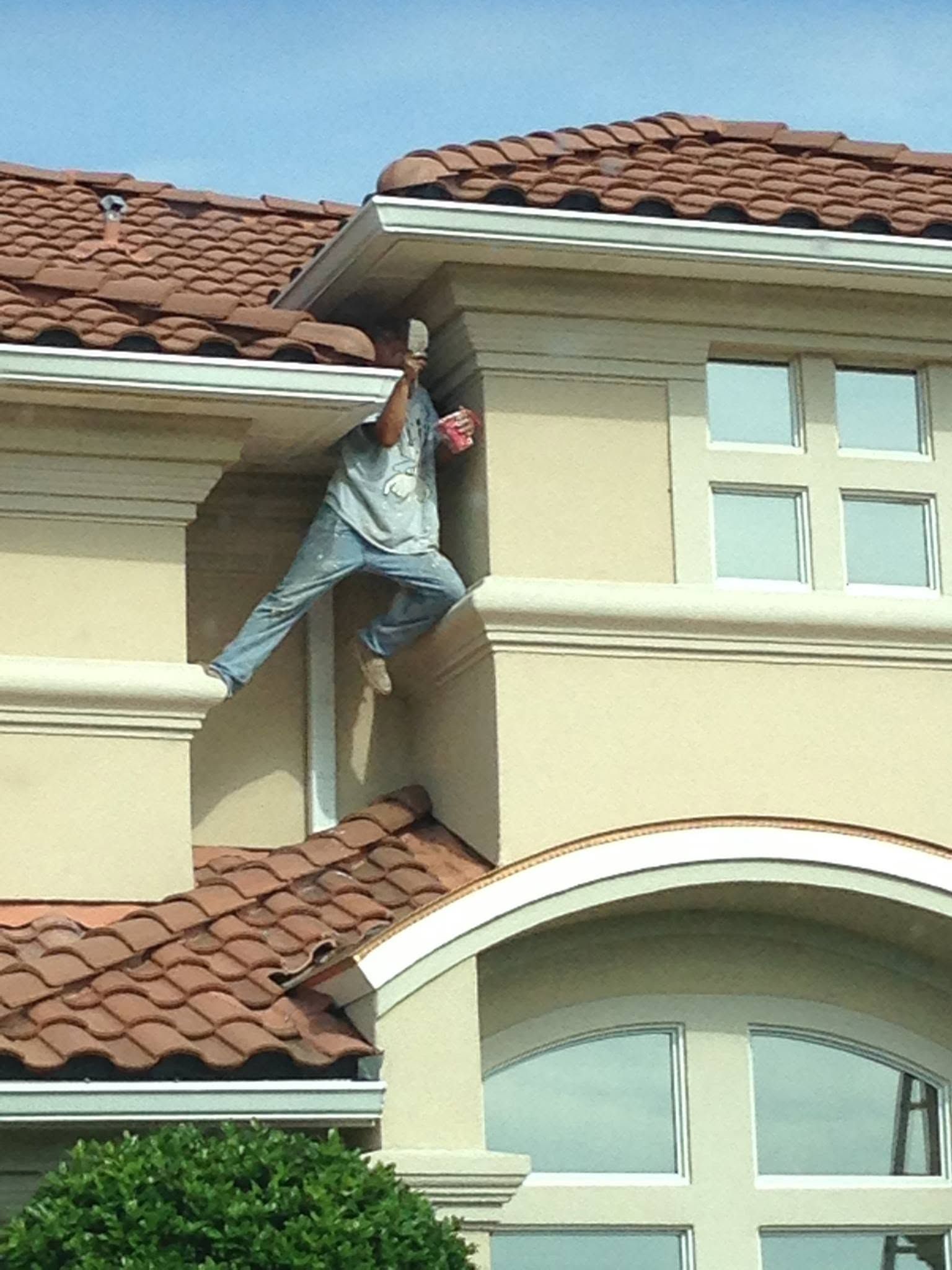 Person hanging from a house roof between two stories, wearing jeans and a gray shirt.