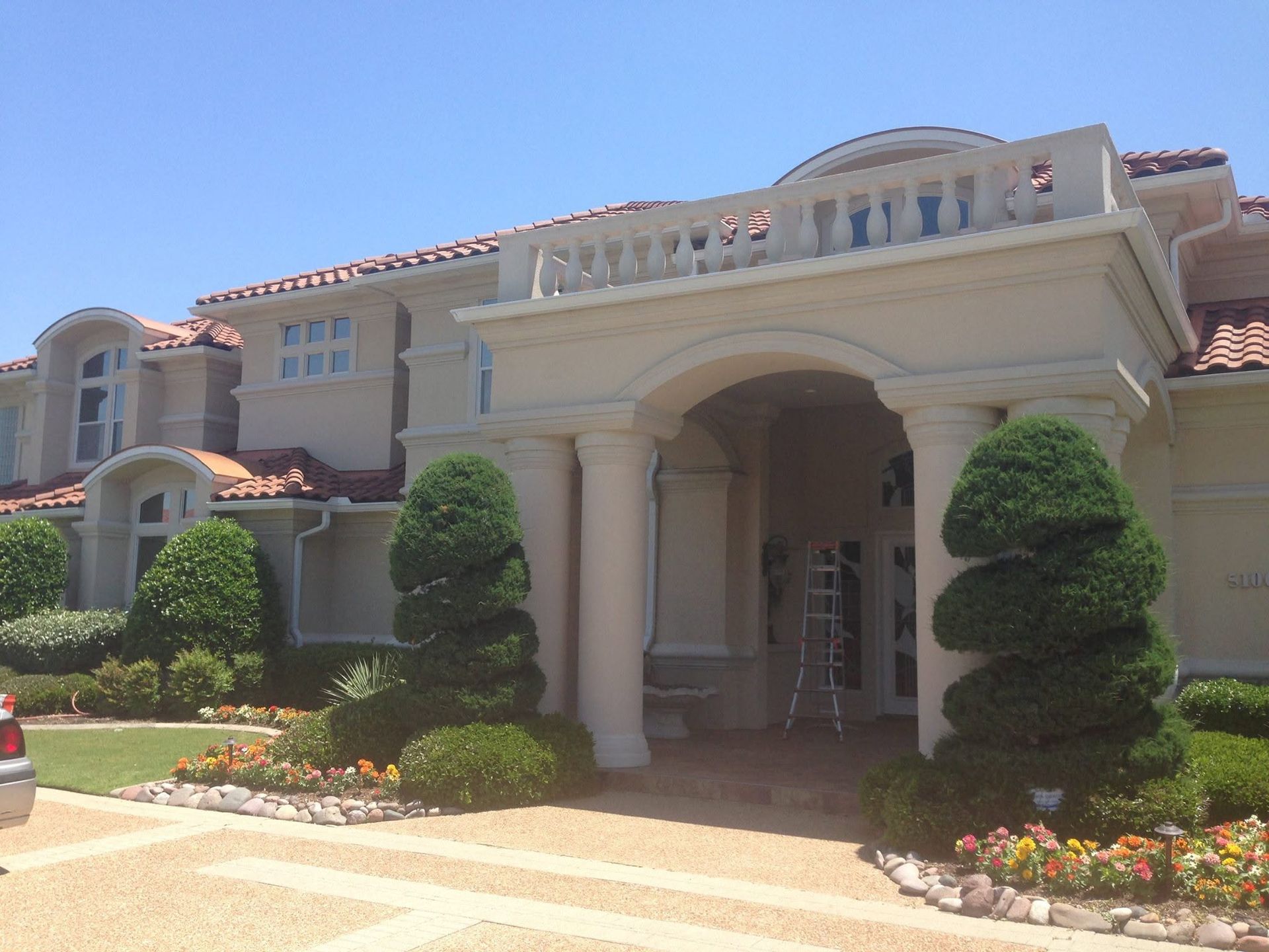 Large beige mansion with red-tile roof, arched entryway, and manicured landscaping under a clear sky