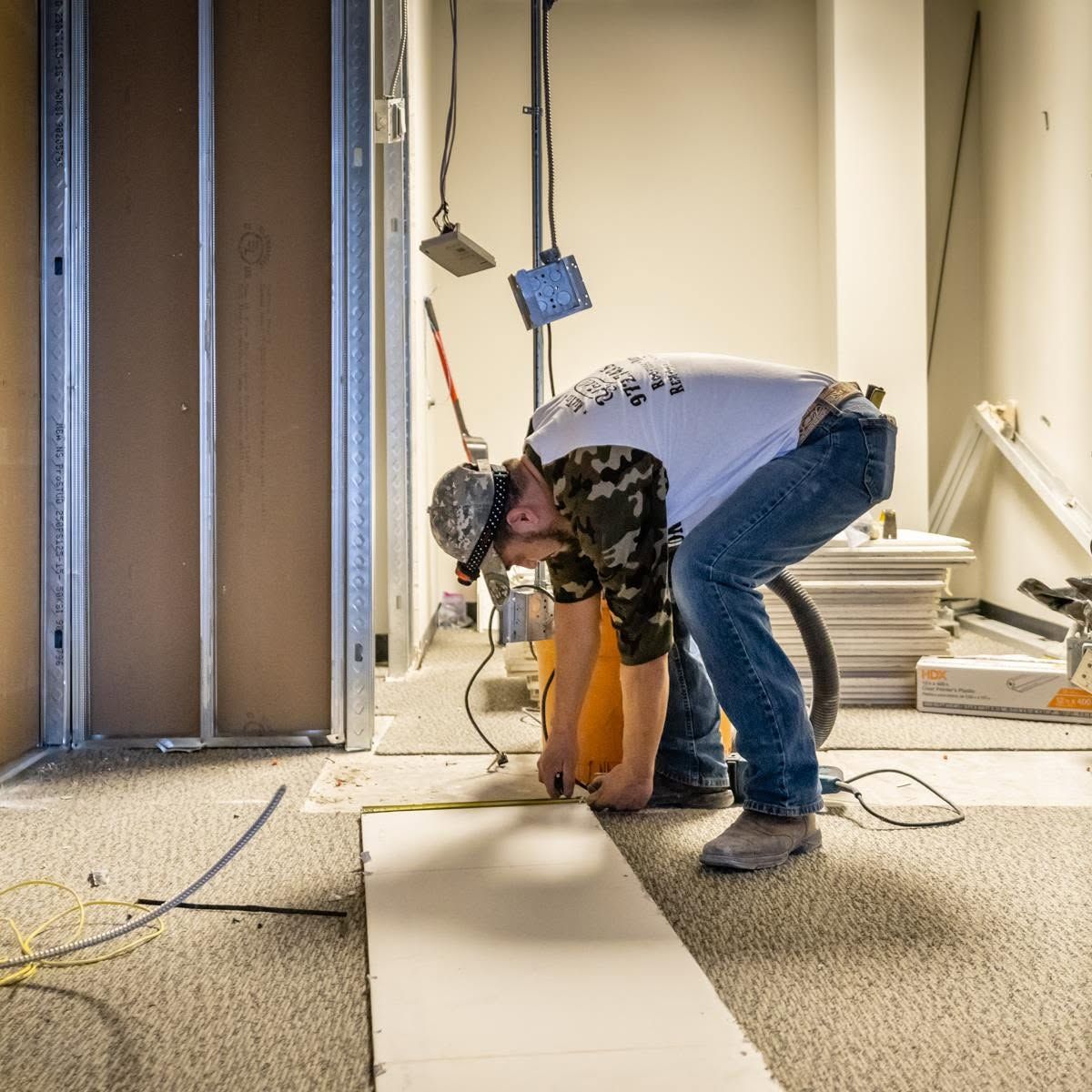 Worker laying flooring in a renovation site, bending over a white panel amid tools and cables.