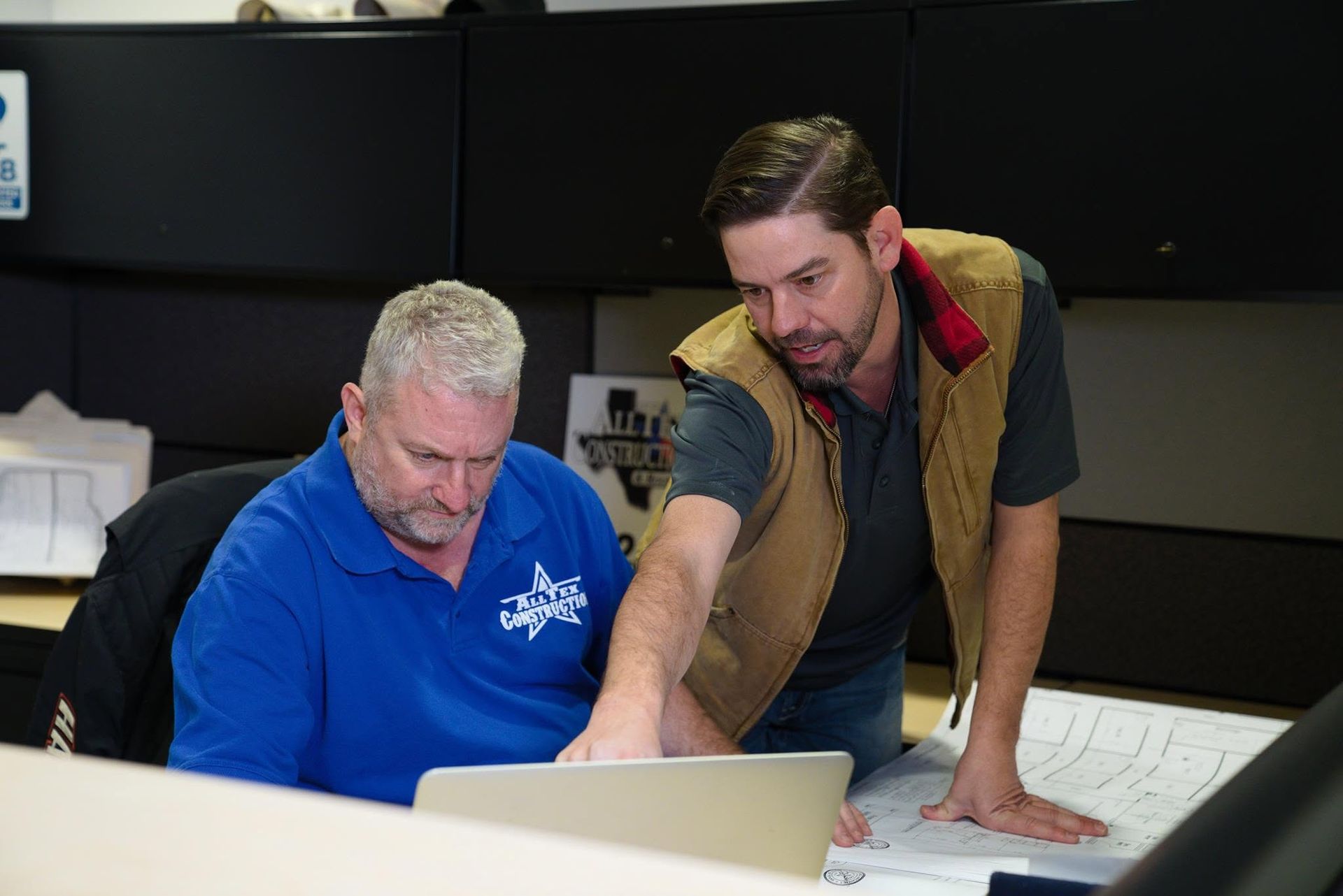 Two coworkers reviewing a laptop in an office, one seated and one leaning over papers.