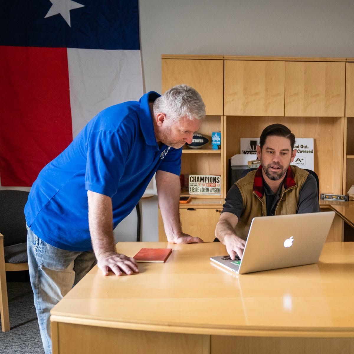 Two men in an office review a laptop at a wooden table, with a flag hanging on the wall.