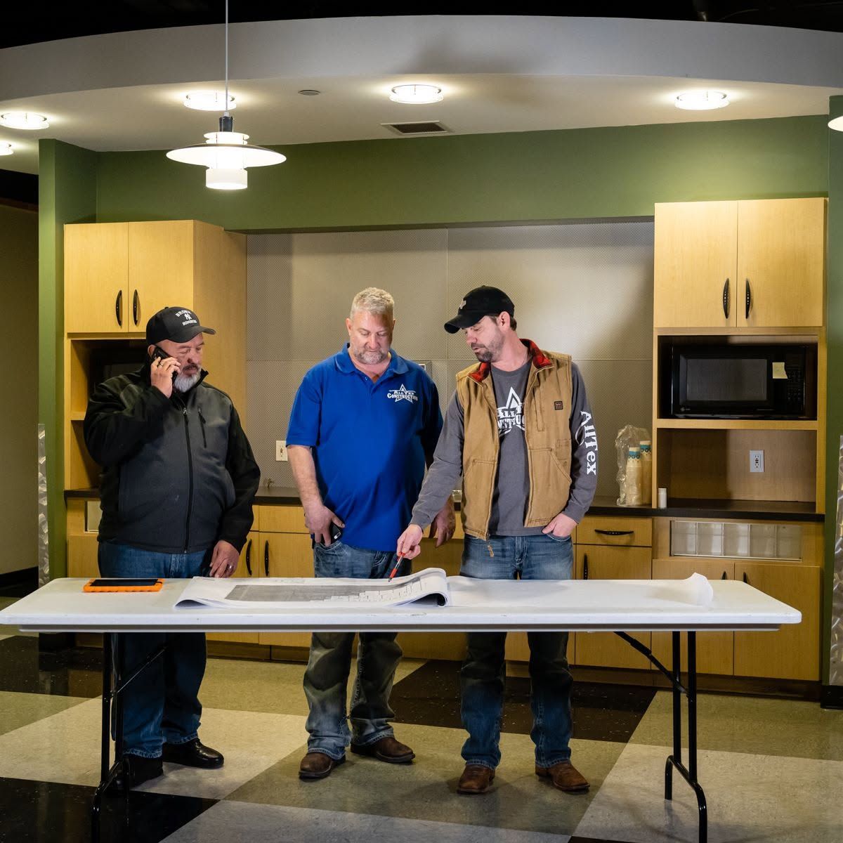 Three men stand behind a folding table in a kitchen, with cabinets, a microwave, and overhead lights.