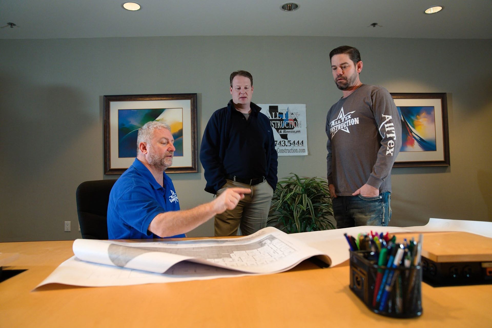 Three people in an office review large blueprints across a conference table.