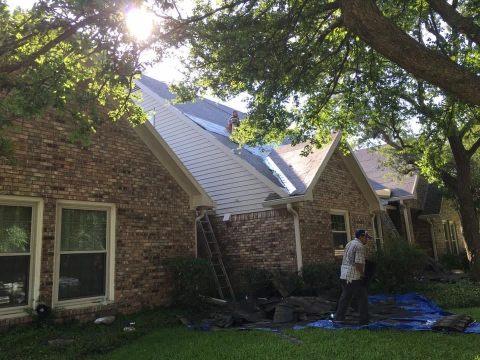Worker on a roof repairing storm damage on a brick house under trees.