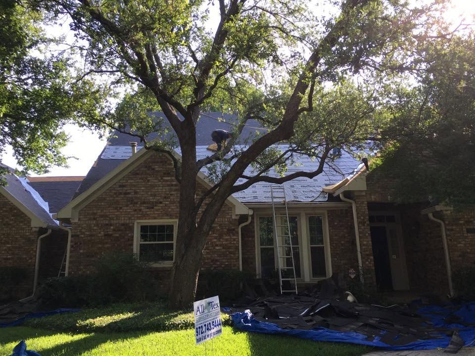 Brick house with a large tree in front and roofing materials on the lawn, suggesting home repair work.