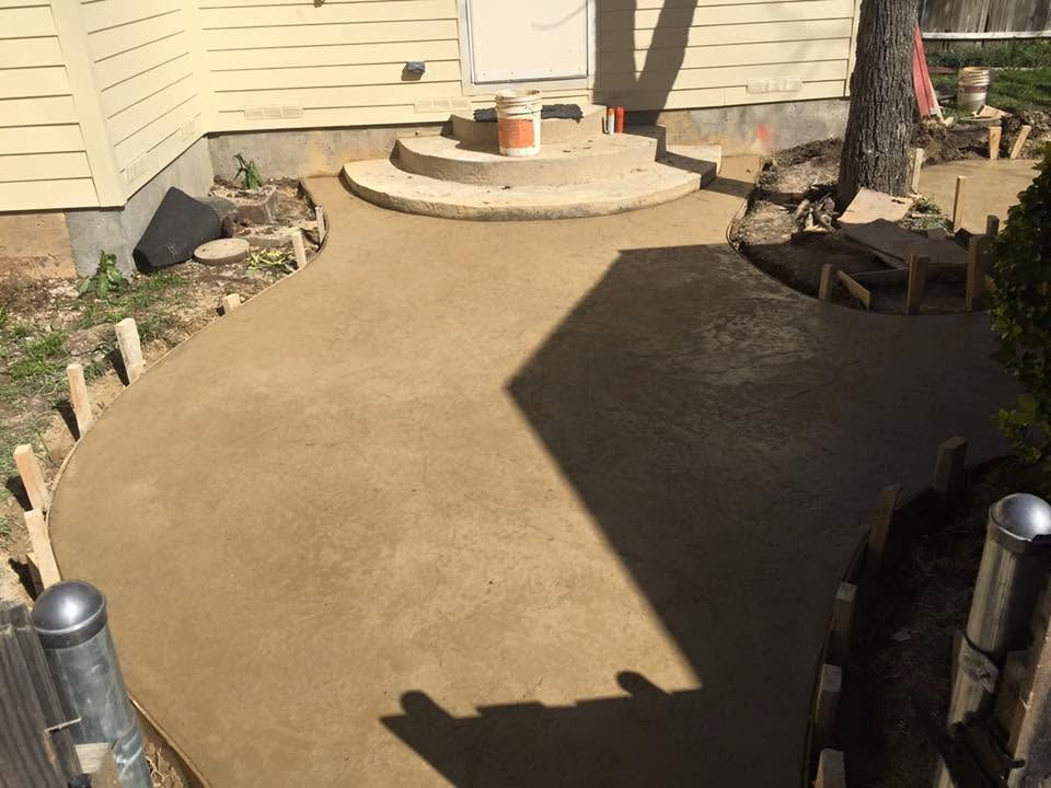 Curved concrete walkway leading to a raised patio with steps and house siding, bordered by shrubs and a tree shadow