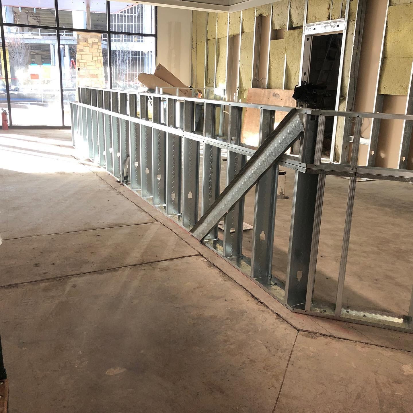 Interior construction site with metal wall framing and a concrete floor near a storefront window