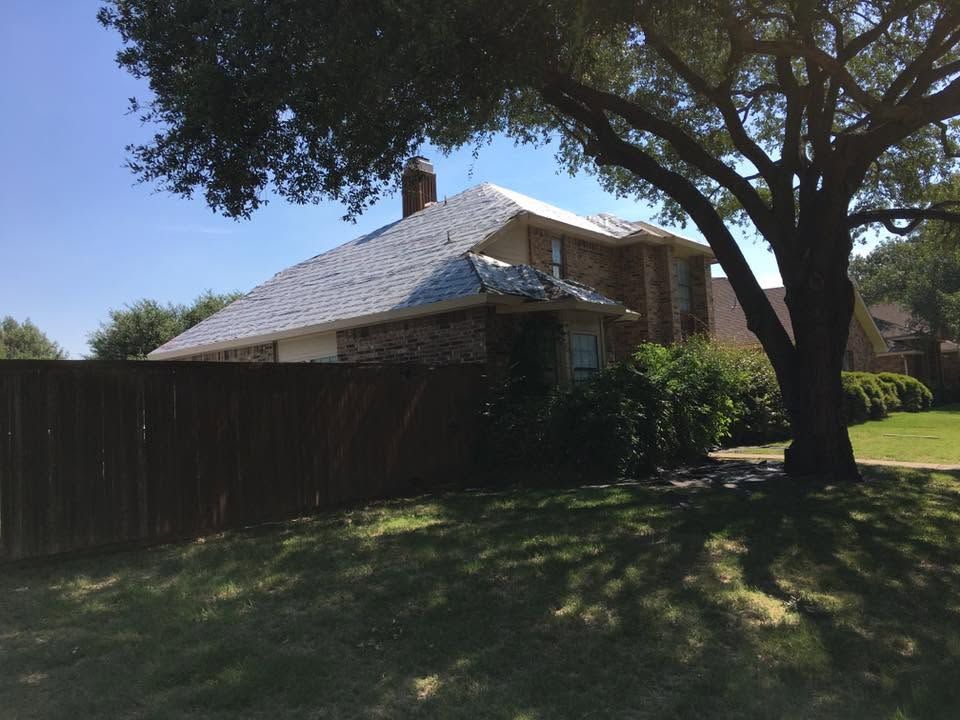 Suburban house with a shingled roof, wooden fence, and large tree casting shade over a grassy yard