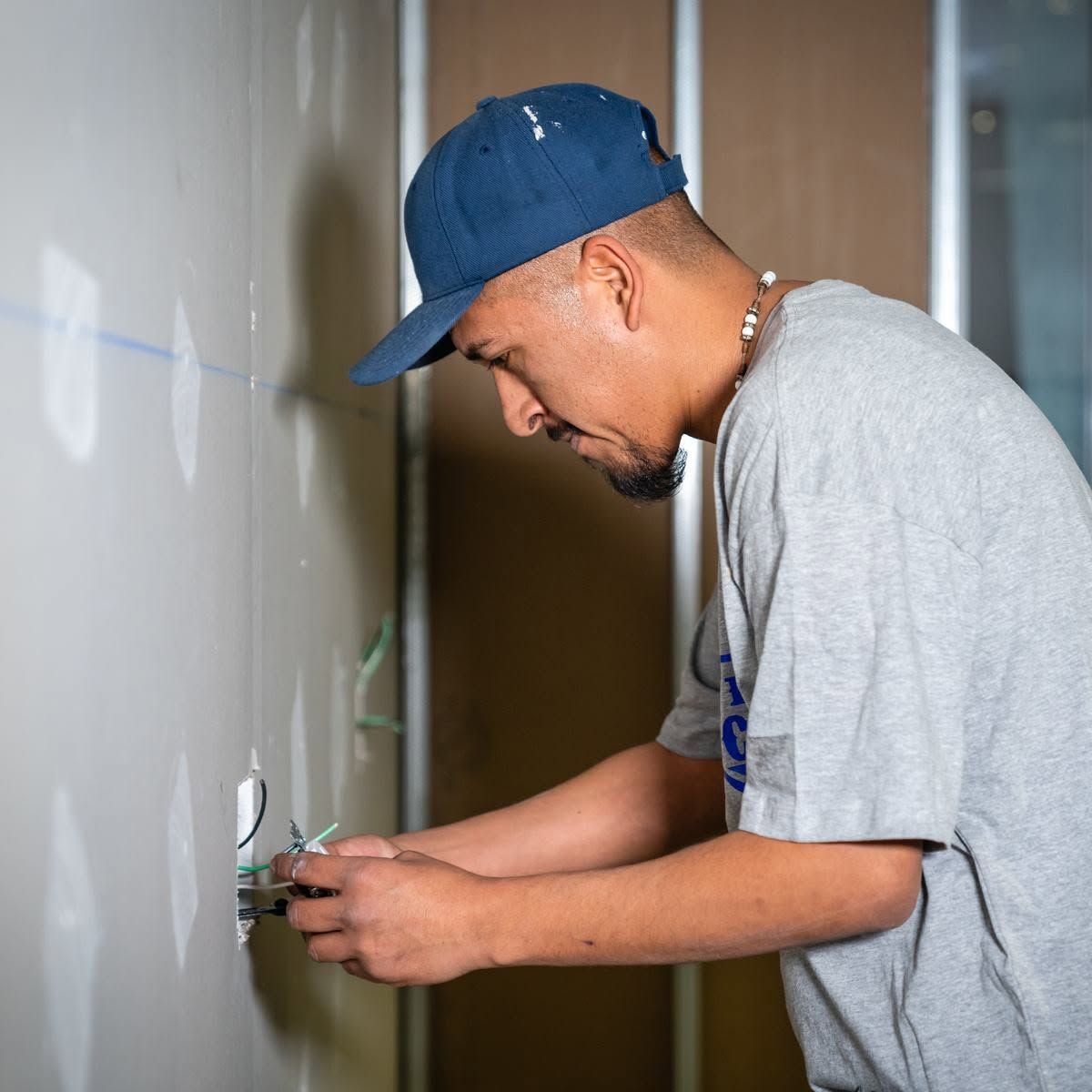 Man installing an electrical outlet in a wall, wearing a blue cap and gray shirt.