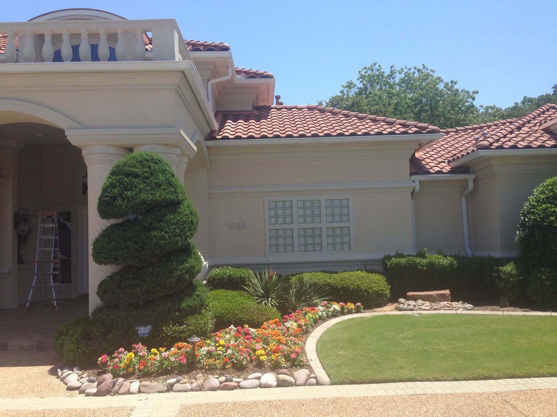 Front entrance of a beige house with red tile roof, manicured lawn, and colorful flower bed