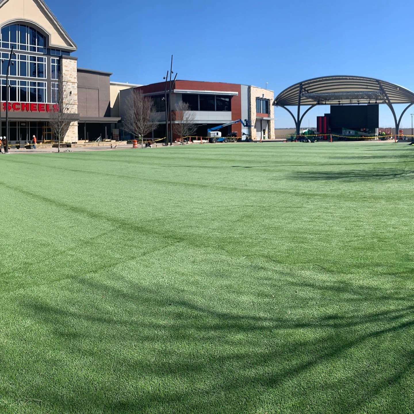 Outdoor shopping center with a large green artificial turf plaza and modern buildings under a clear sky