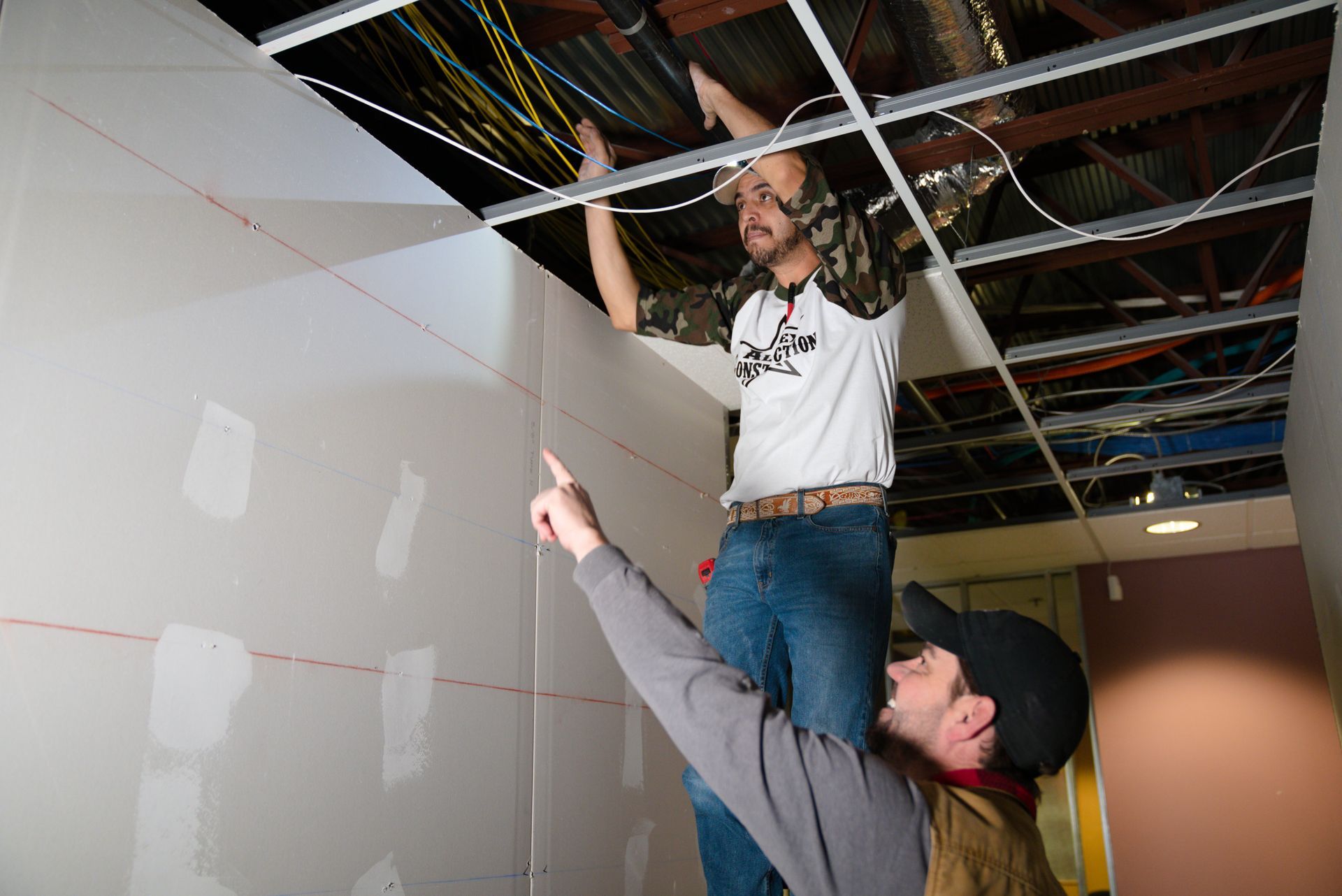 Two people working in a construction site, one holding another up near a ceiling with exposed wiring.
