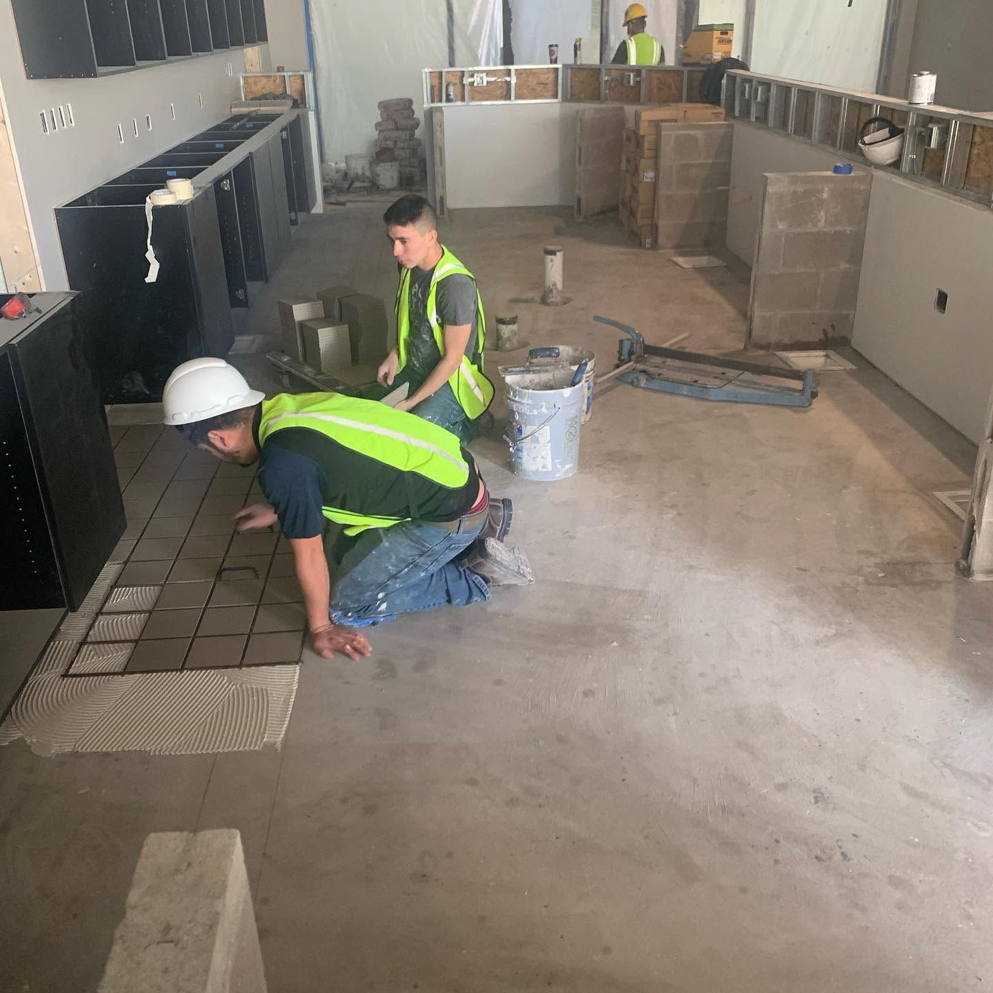 Two workers in neon vests and hard hats kneel on a tiled floor during indoor construction.