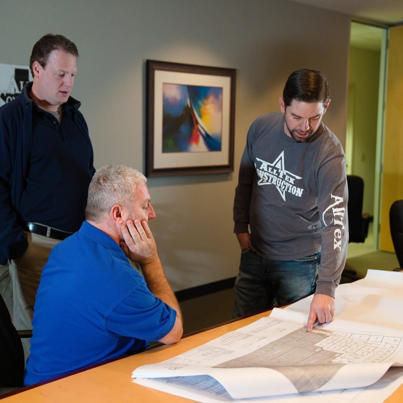 Three people review large blueprints at a table in an office, one pointing while the others look on.