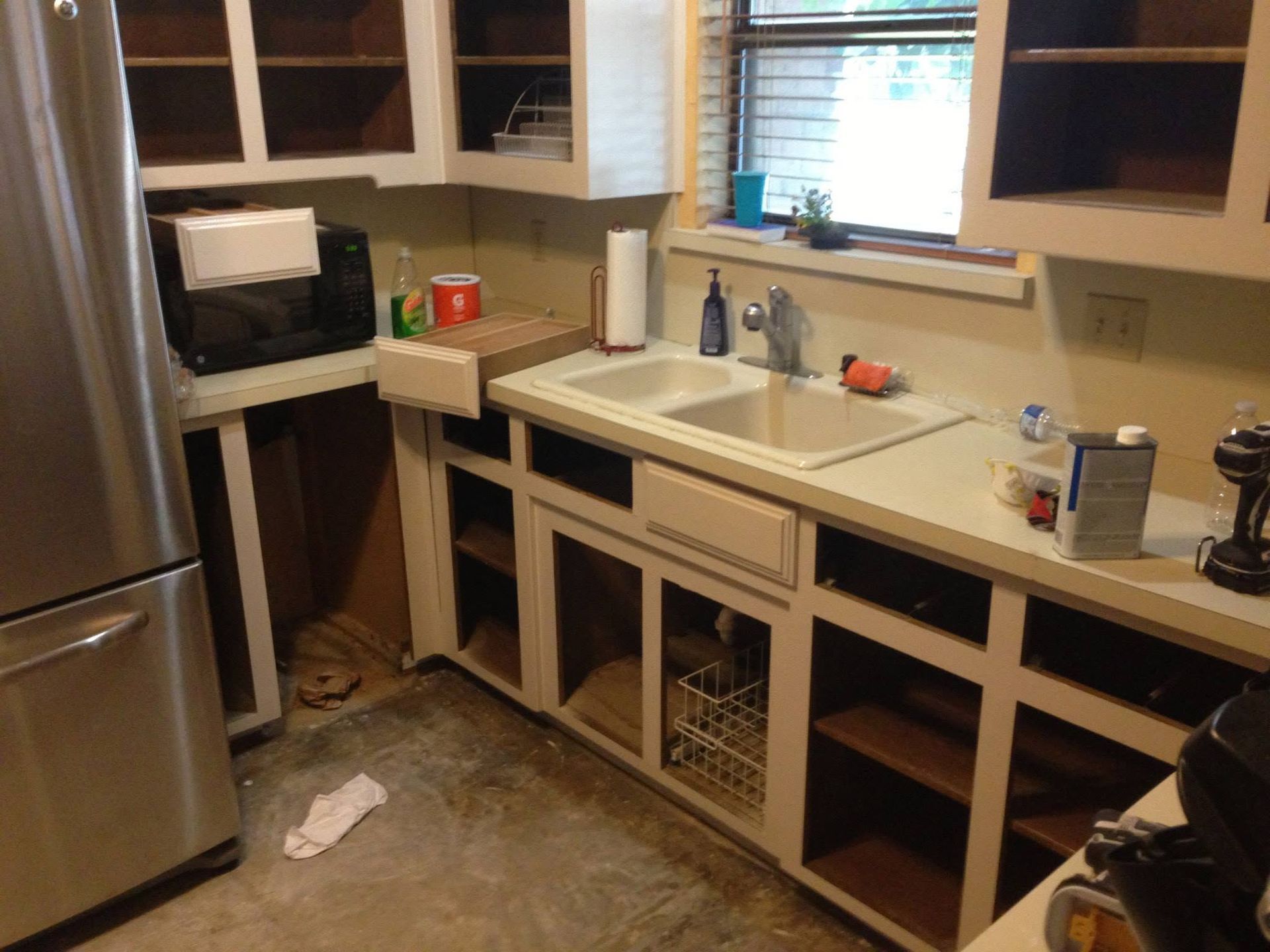 Kitchen with unfinished cabinets, stainless fridge, sink under window, and cluttered floor.