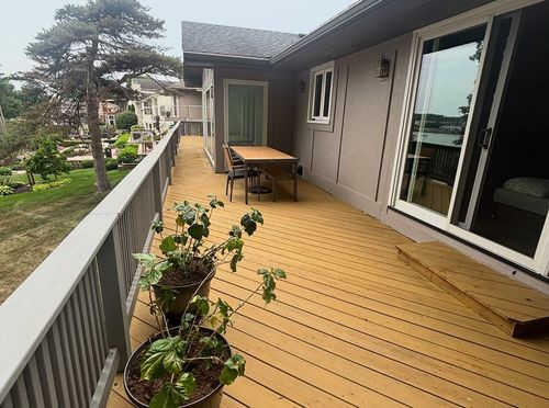 Wooden balcony with potted plants, a table and chairs, overlooking a green yard and trees.