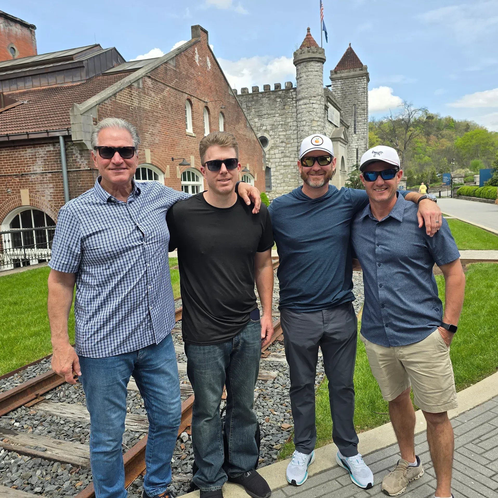 Four men standing together outdoors by a historic stone building and castle wall, smiling in casual clothes
