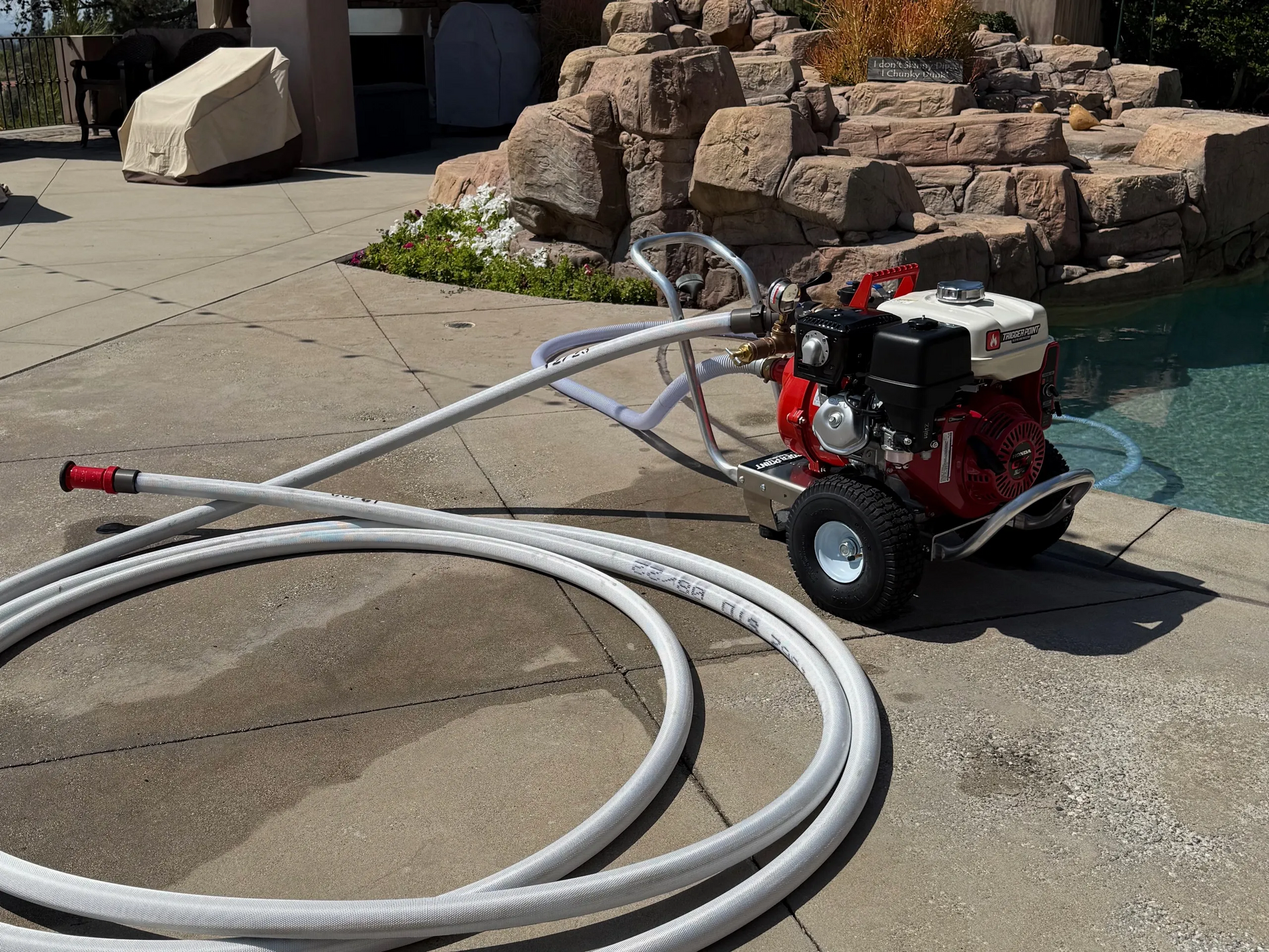 A red and black pump with white hose near a pool and rock formation.
