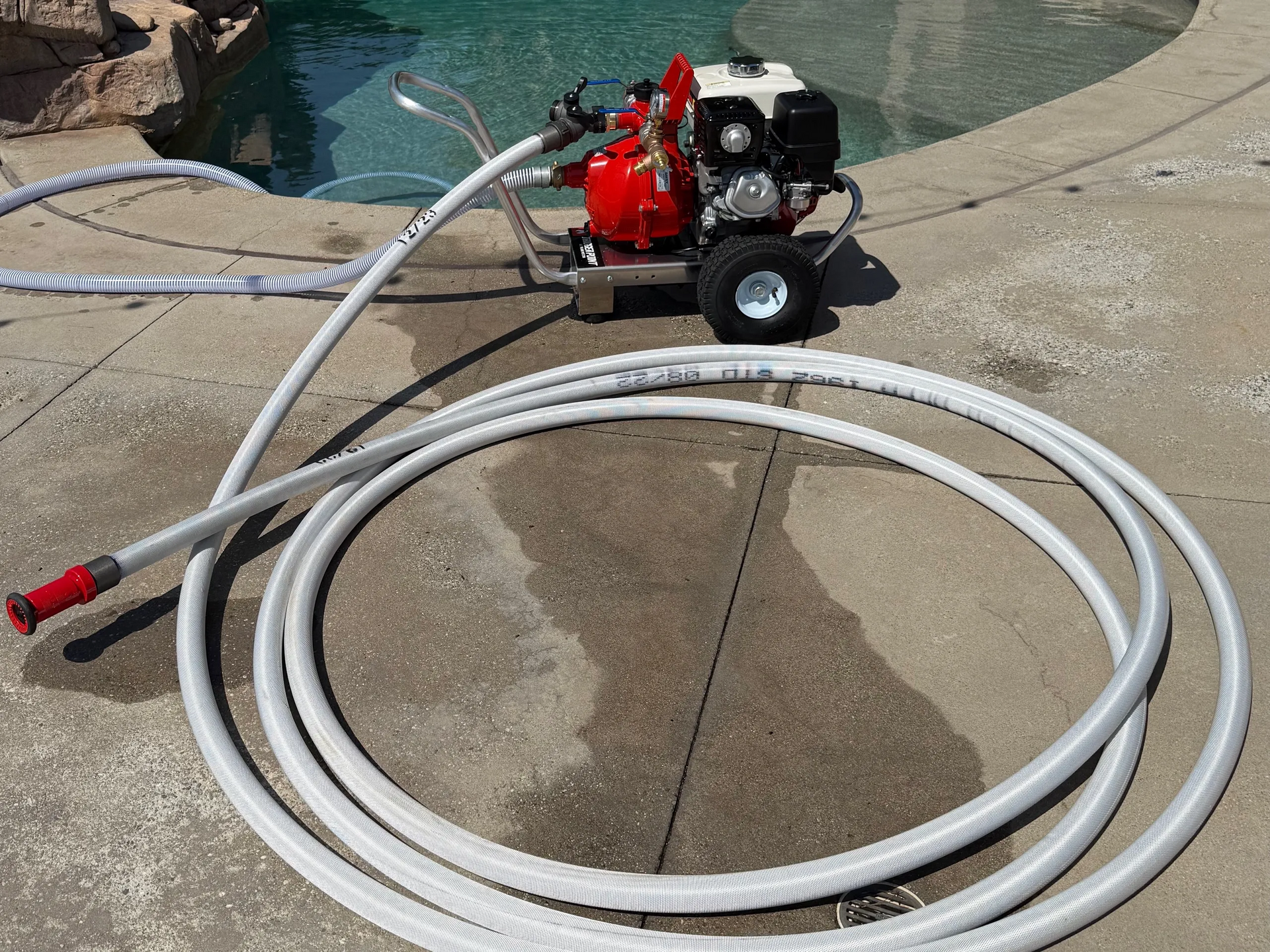 Gas-powered water pump near a pool, with white hoses laid out on a concrete surface.