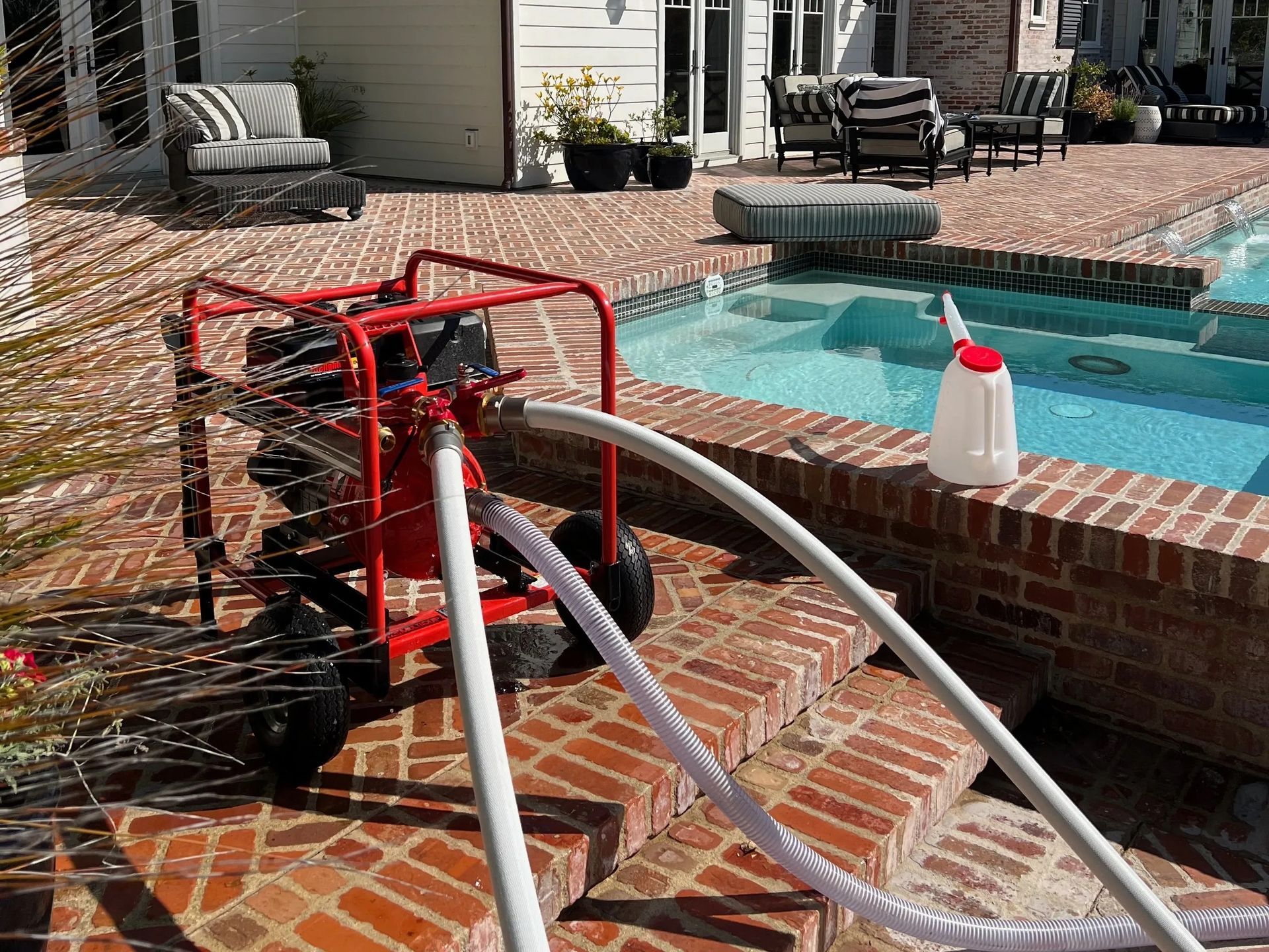 A red pump is draining a swimming pool. Hoses and a white container are present on the brick patio.