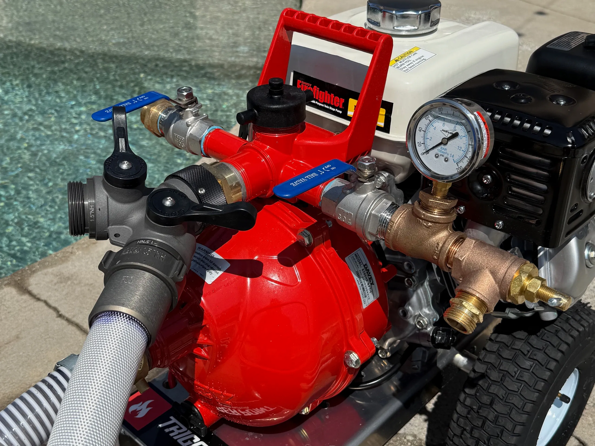 Red water pump with hoses, valves, and gauge; engine on a wheeled base near a pool.