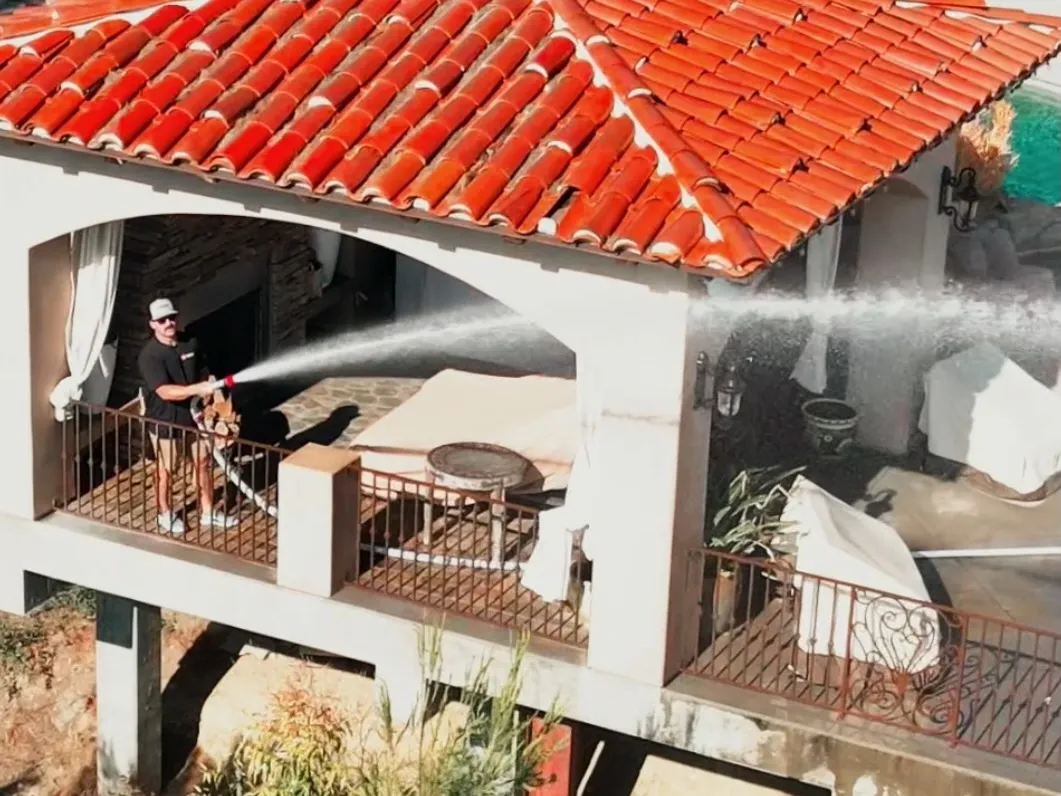 Man washing balcony with hose; Spanish-style building with red tile roof.