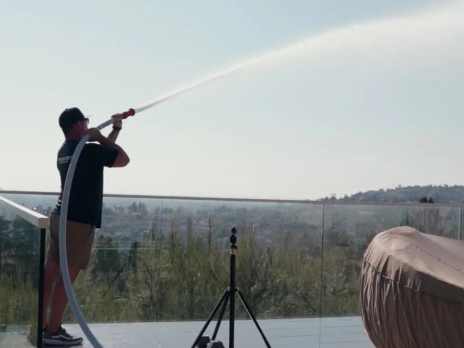 Man spraying water with a hose on a sunny day from a rooftop overlooking a city.