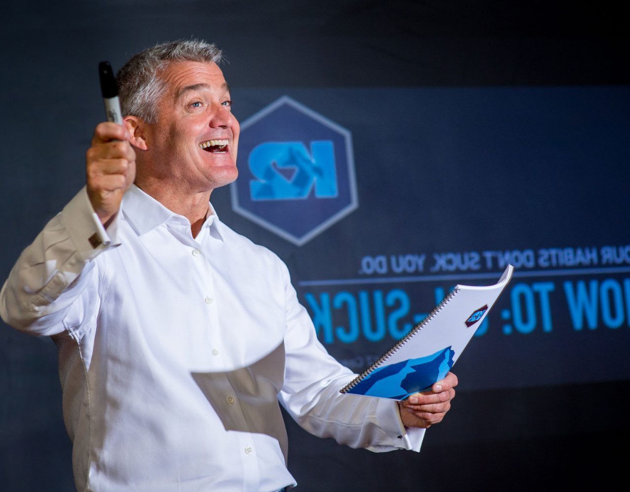 kris kaplan in a white shirt is holding a notebook and pointing