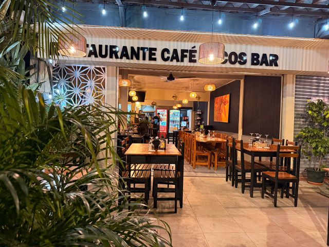 Outdoor café entrance with wooden tables, warm lights, and greenery at dusk, restaurant las gemelas Paco bar Manuel Antonio