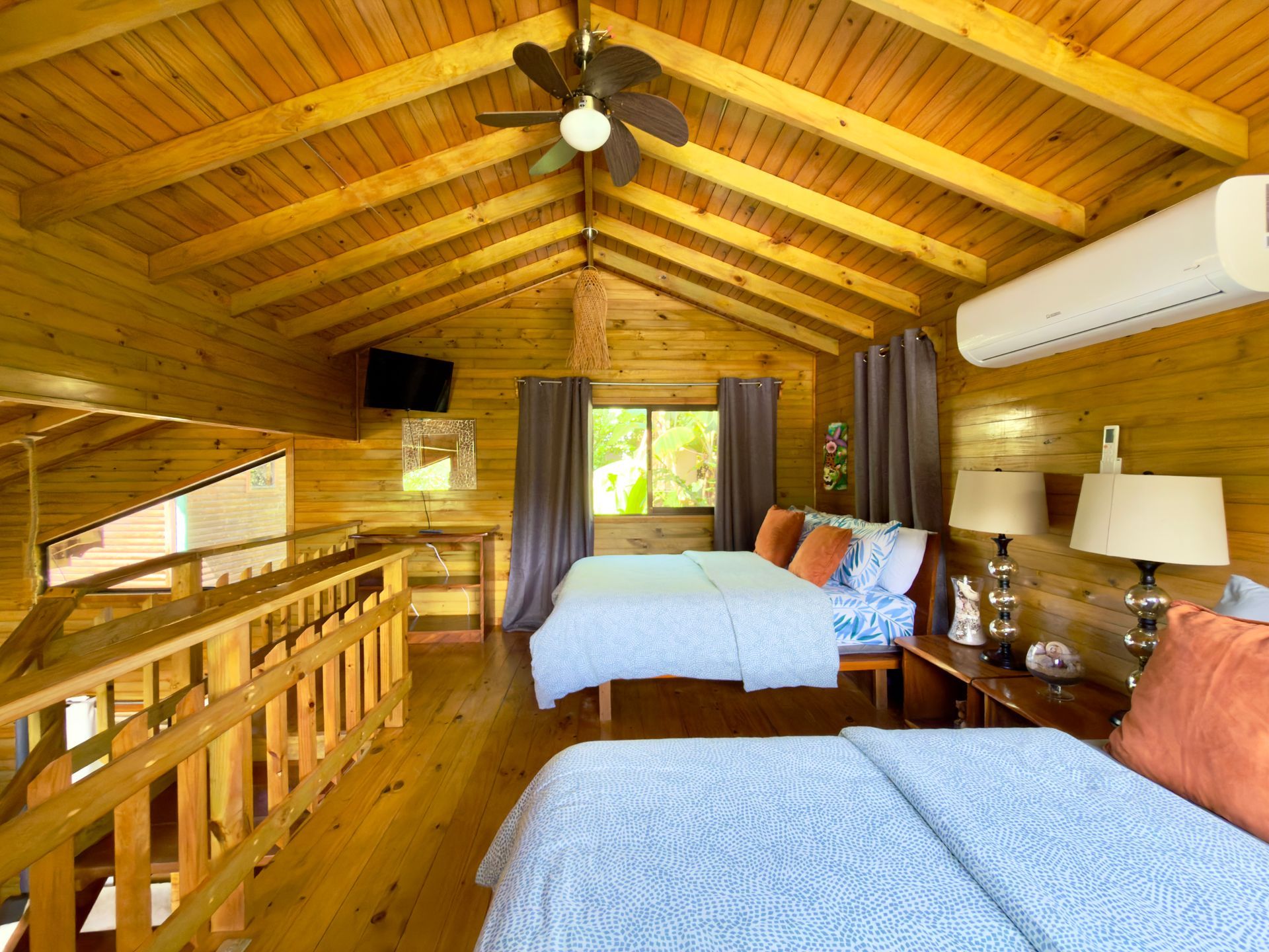 Cozy wood-paneled loft bedroom with two beds, ceiling fan, and large window under a vaulted ceiling, with air conditioning