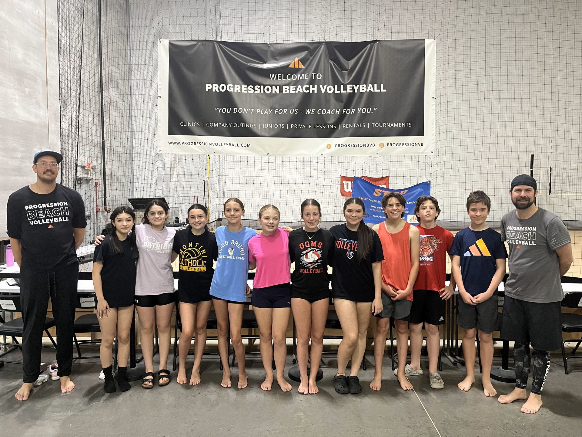 Volleyball team group photo in a gym, standing in front of a banner, wearing matching black jerseys