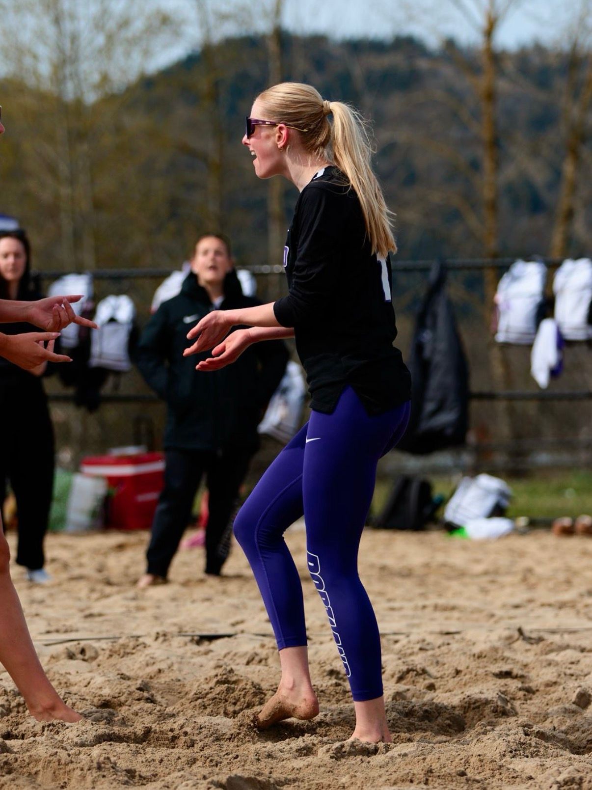 Two beach volleyball players jump at the net to block a volleyball against an opponent on a sunny, outdoor sand court.