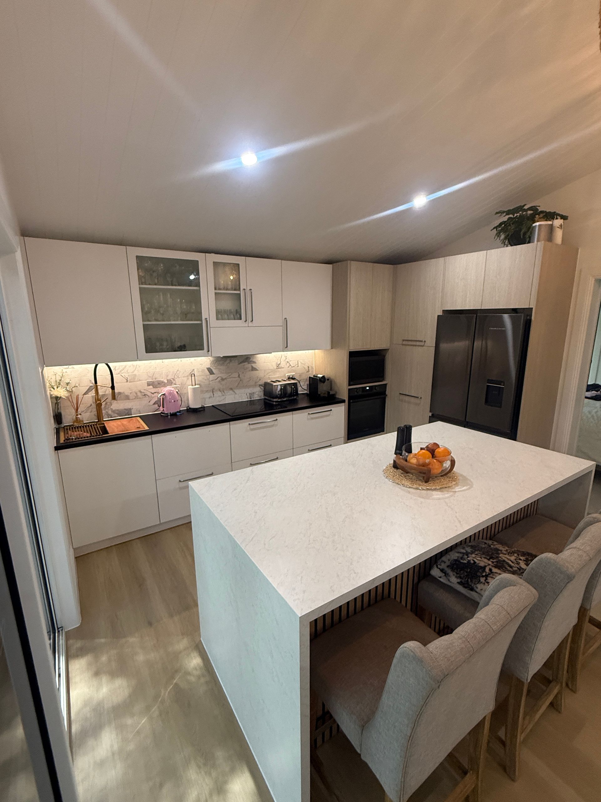 Modern Kitchen with White Cabinets, Black Refrigerator, Island Table, and Beige Chairs — JPR Concepts in Burleigh Heads, QLD