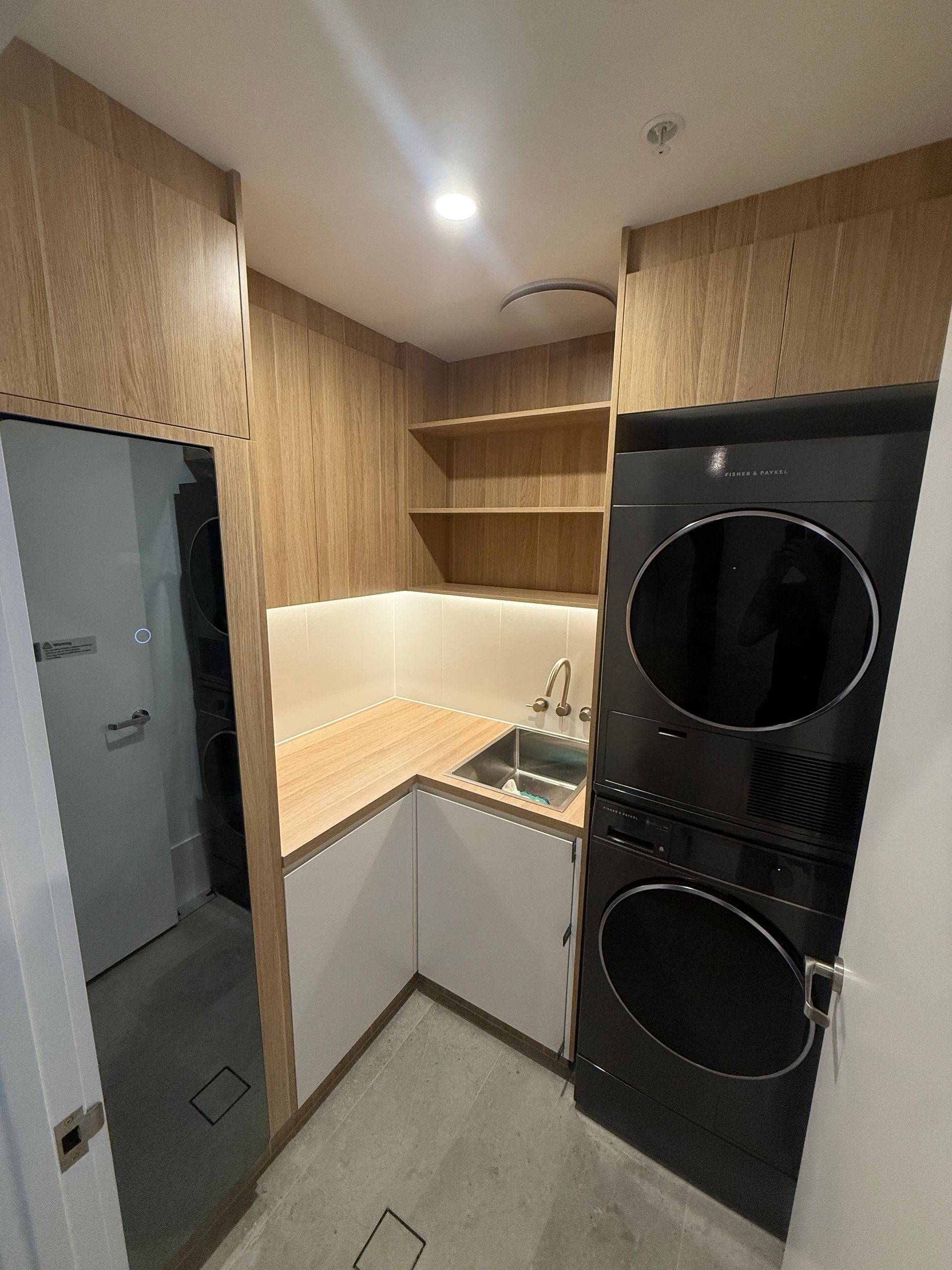 Small Laundry Room with Wood Cabinets, Beige Countertop, Sink, and Stacked Washer and Dryer — JPR Concepts in Burleigh Heads, QLD