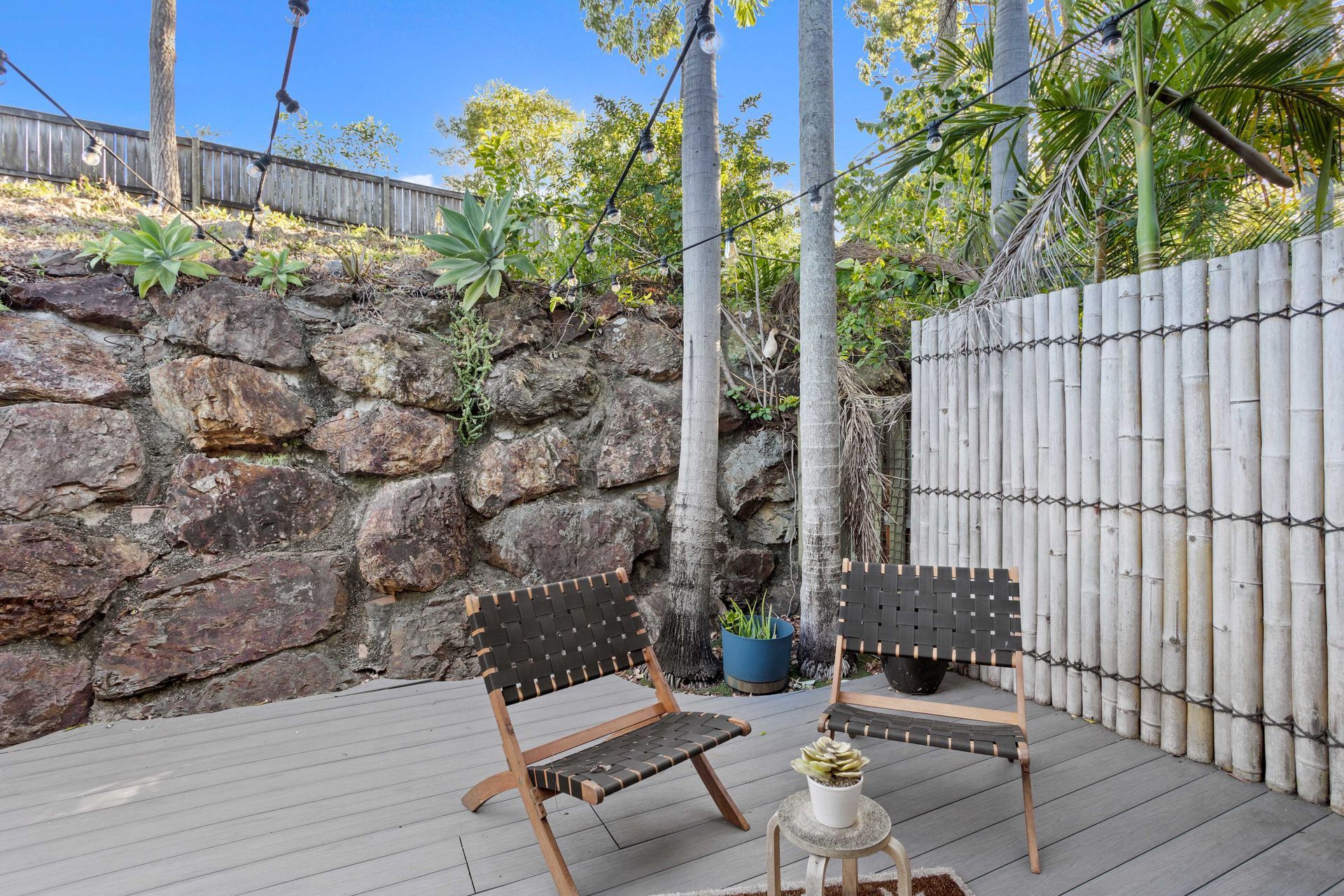 Small Patio with Wooden Chairs and Potted Plants Beside a Stone Wall and Bamboo Fence — JPR Concepts in Burleigh Heads, QLD