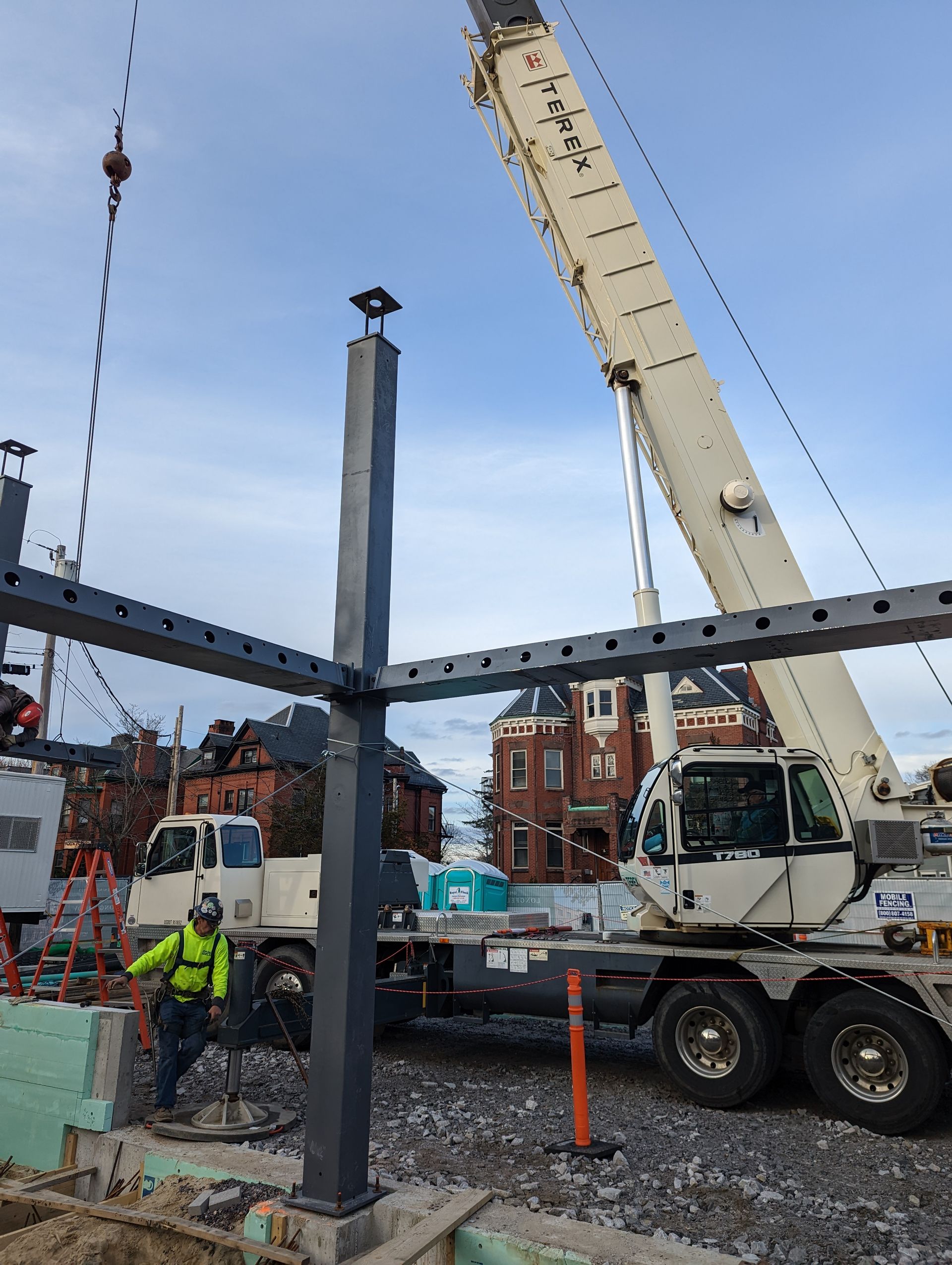 Construction site with crane lifting a steel beam above workers and trucks on muddy ground.