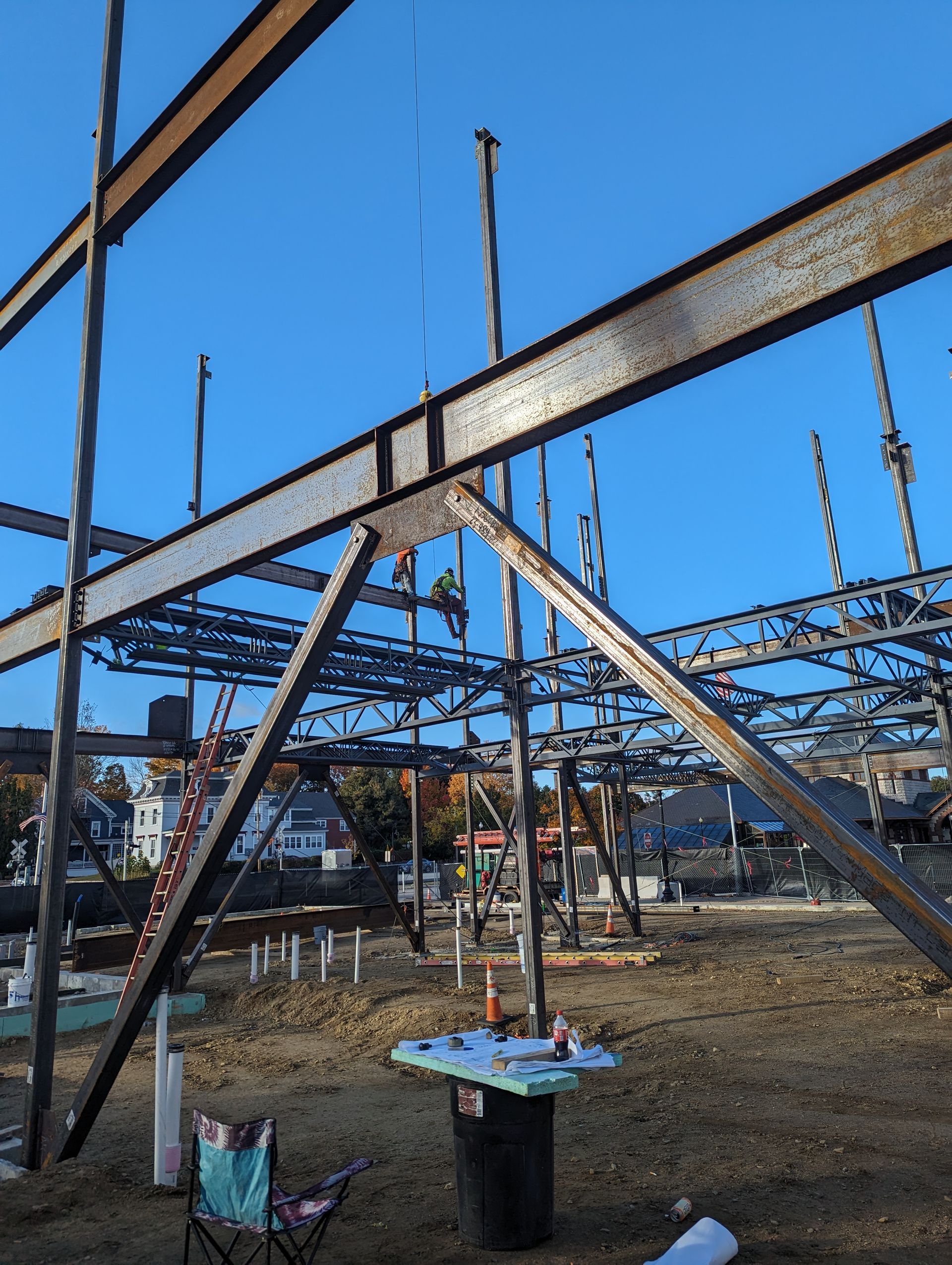 Steel bridge framework under construction against a blue sky, with beams, cables, and a small work table below.