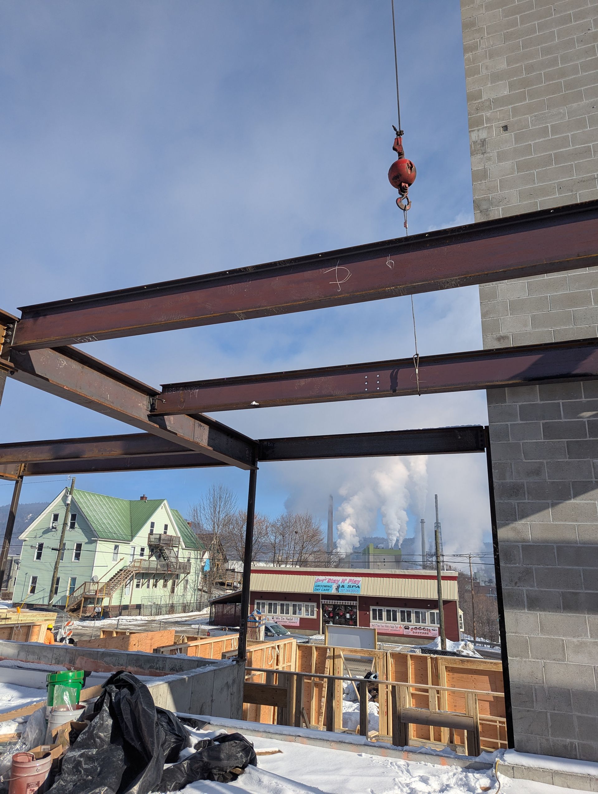 Construction site with steel beams and crane hook against a blue sky, near buildings and snow-covered ground