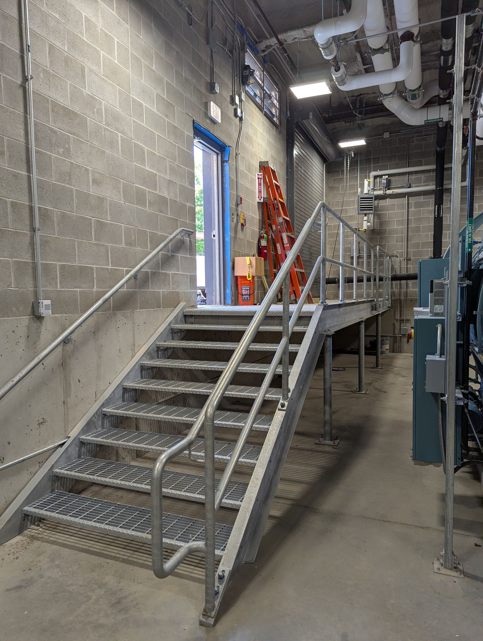 Metal stairs and landing in an industrial utility corridor with cinderblock walls and pipes overhead