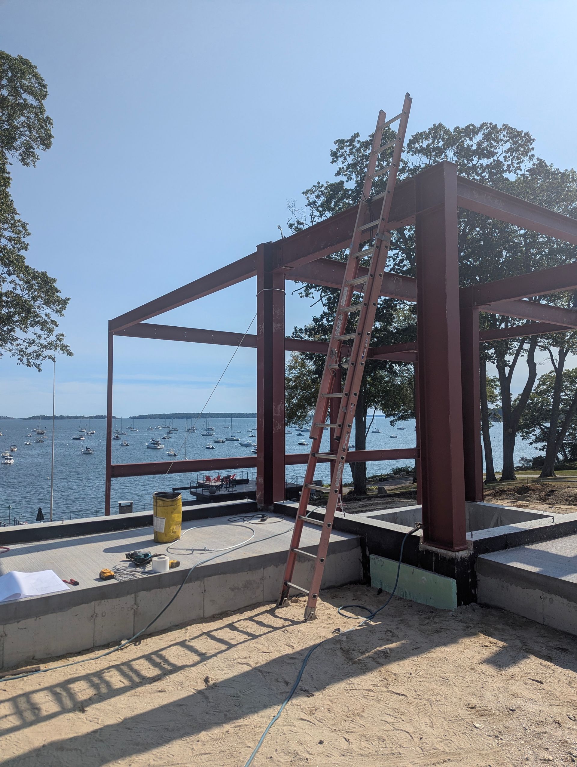 Wooden pergola under construction by a waterfront, with a ladder leaning against it and the sea in the background