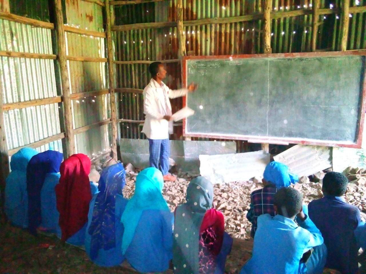Teacher in white coat points to a chalkboard in a small classroom; students in blue sit on the ground, some wearing head coverings.