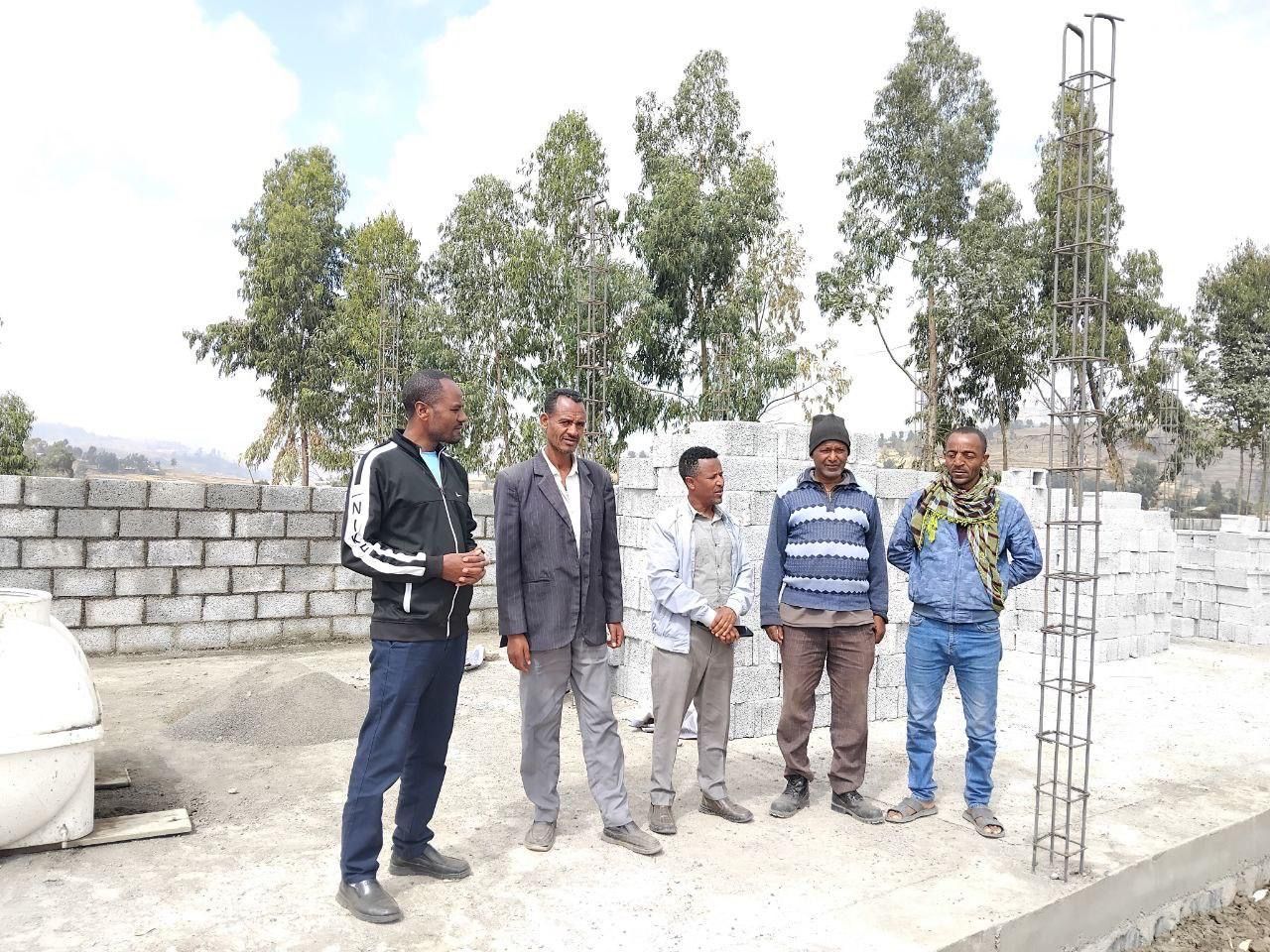 Five men standing near a construction site with concrete blocks and a steel frame. Outdoors, daytime.