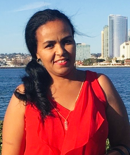 Woman in red top smiles outdoors, with city skyline and water backdrop.