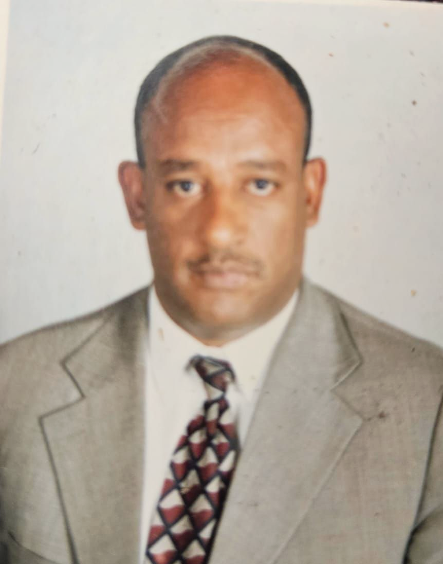 Man in suit and tie, brown skin, looking at the camera. Neutral expression, indoor setting.