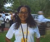 Woman with glasses smiles, wearing white shirt, black lanyard, standing outside near cars.